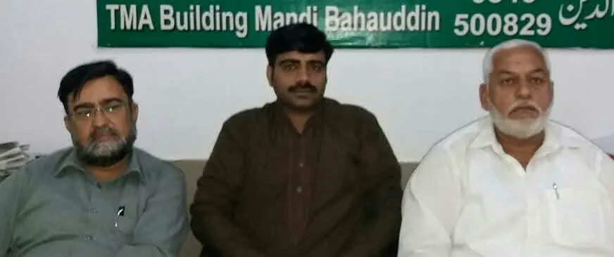 Three men standing outside a building with a TMA Mandi Bahauddin sign.