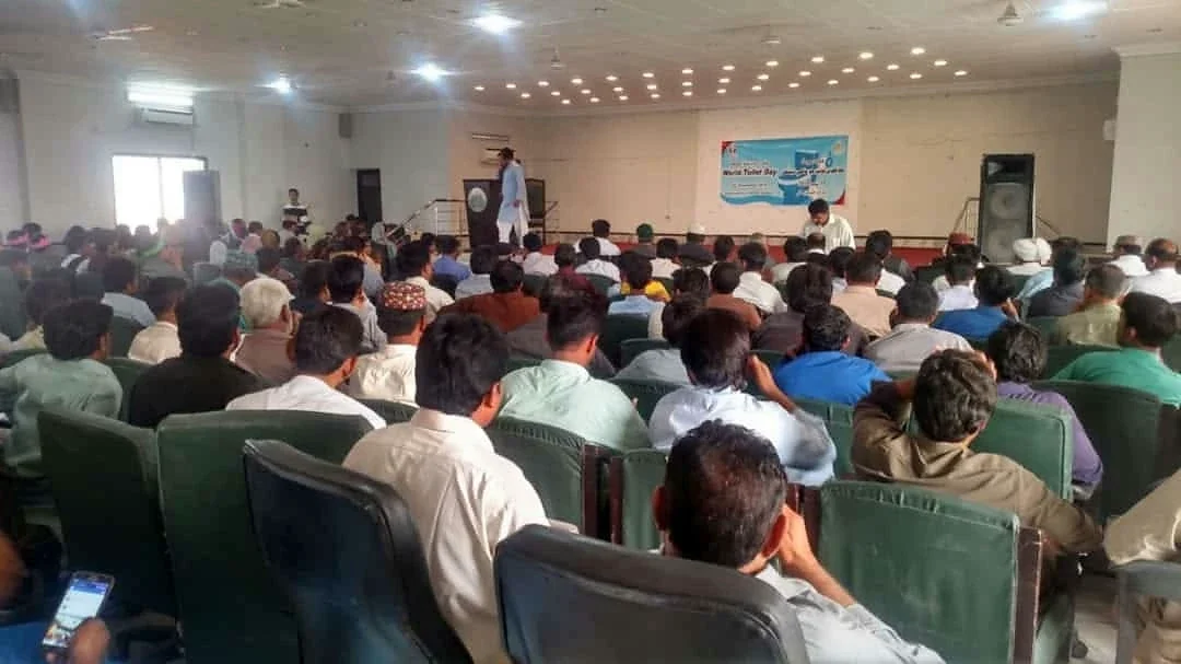 An audience facing a stage with a speaker and a blue backdrop.