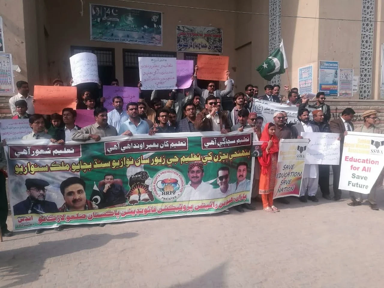 A group holding Urdu banners and coloured placards outside a building.