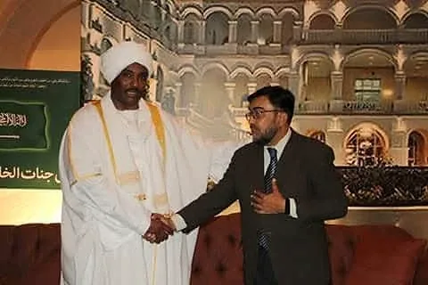 A man in a suit shaking hands with a man in white robes at an indoor event.