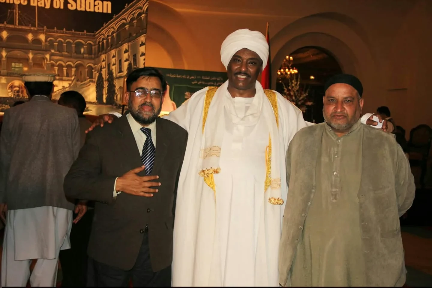 Three men posing at a reception in front of a printed event backdrop.