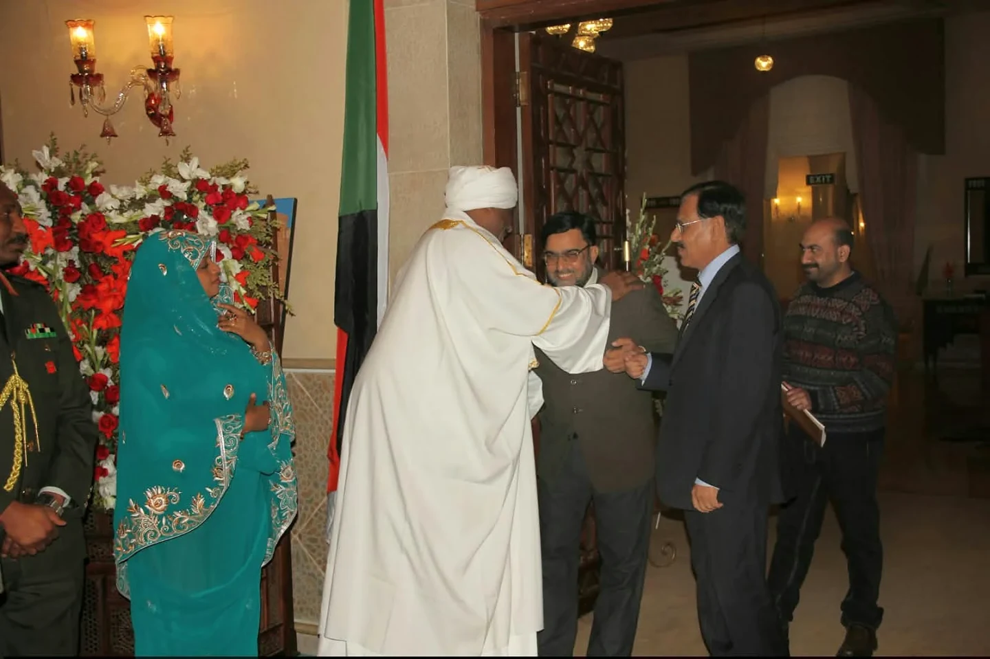 Two men shaking hands indoors beside a tall floral arrangement.