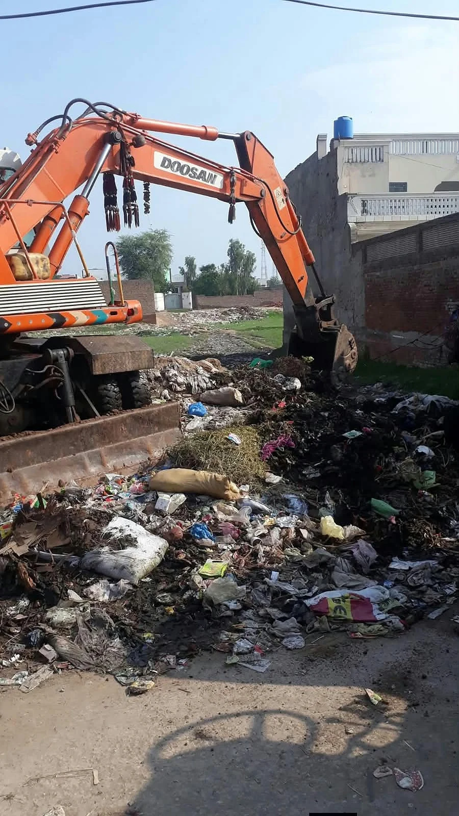 An orange excavator beside a channel and a pile of waste near buildings.