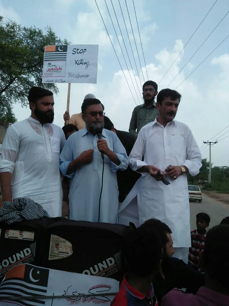 A man speaking into a microphone beside people holding flag-themed placards.