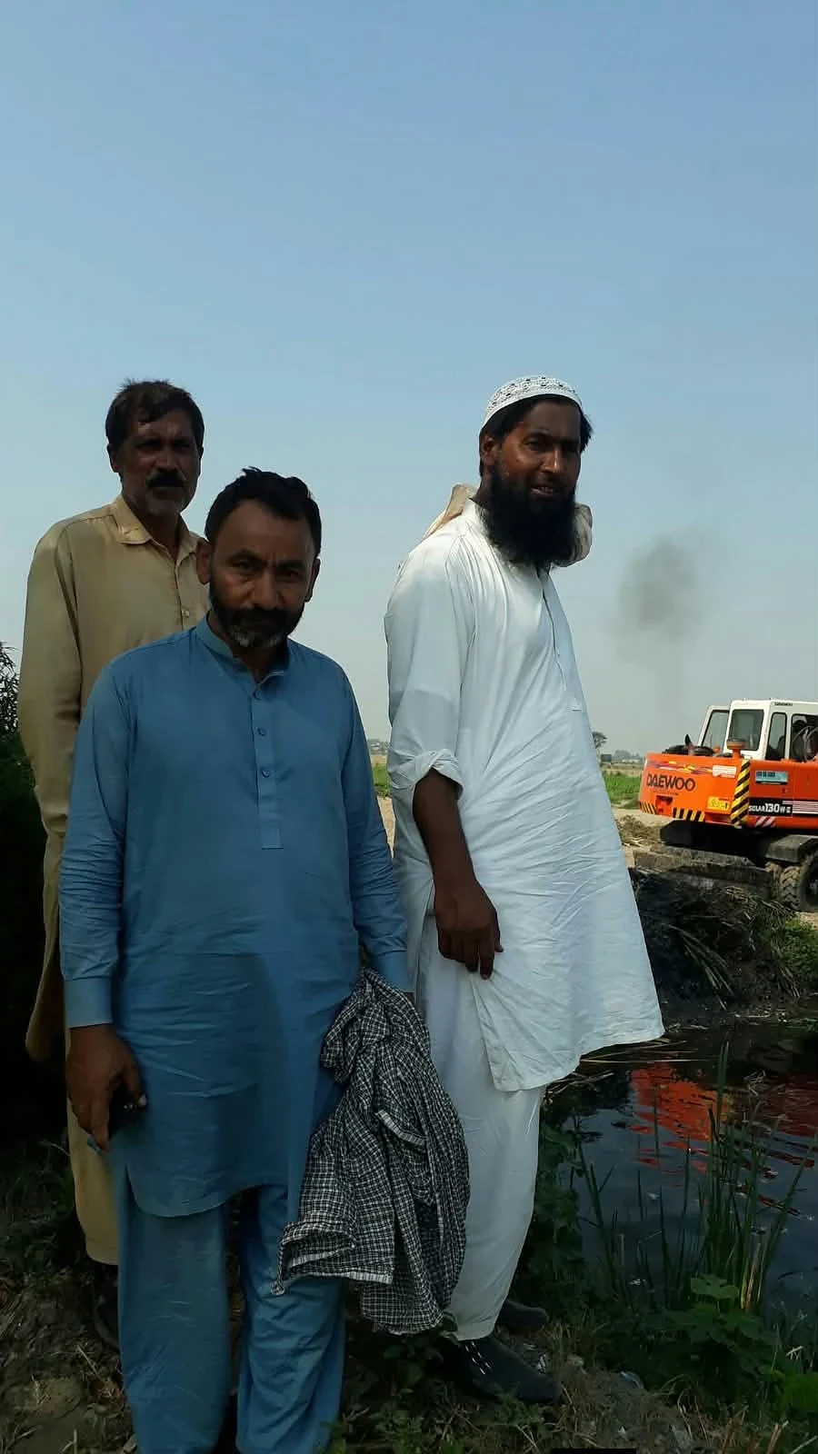 Three men standing near vegetation and machinery outdoors.