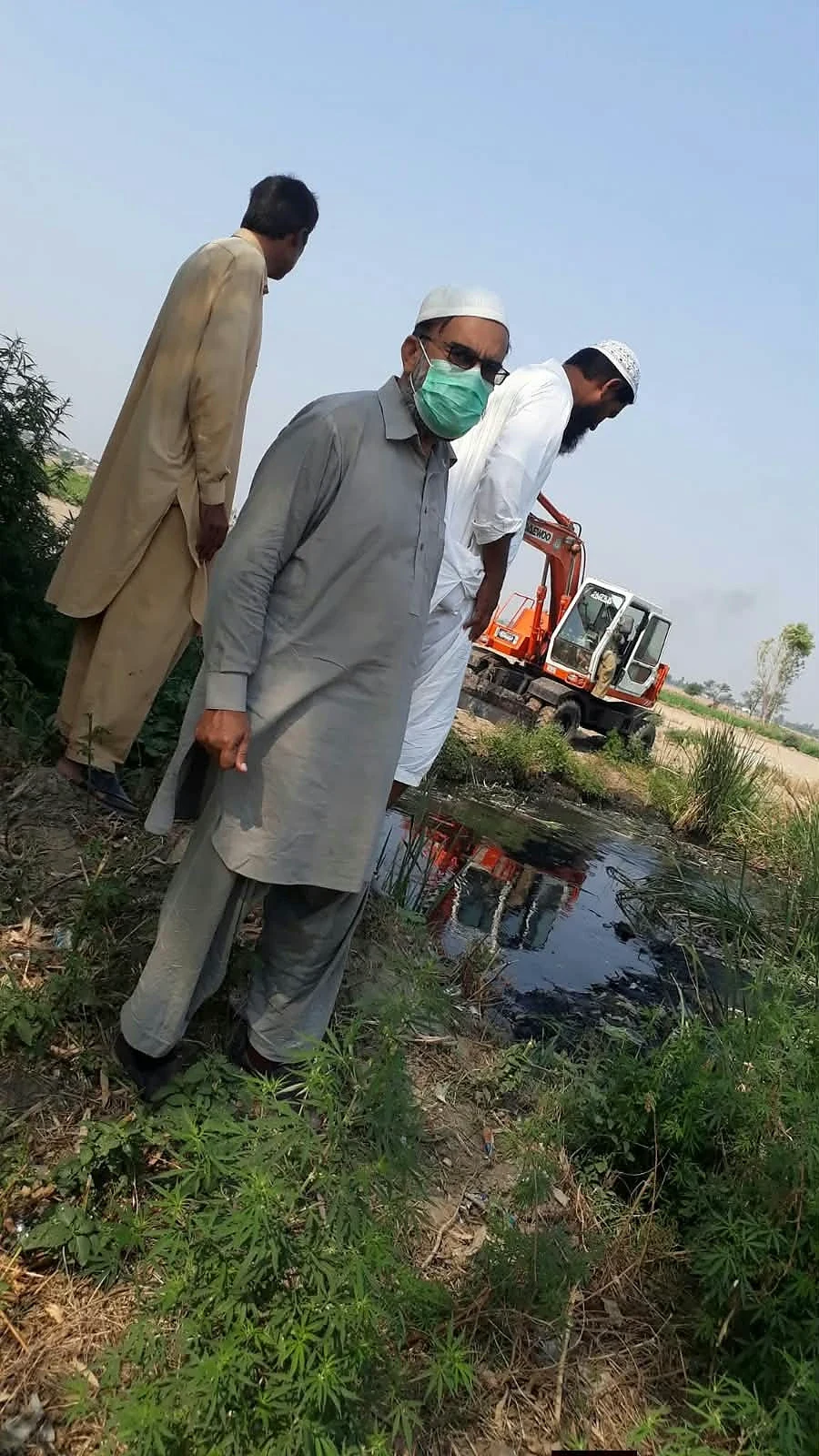 Three men standing beside a narrow channel and vegetation.