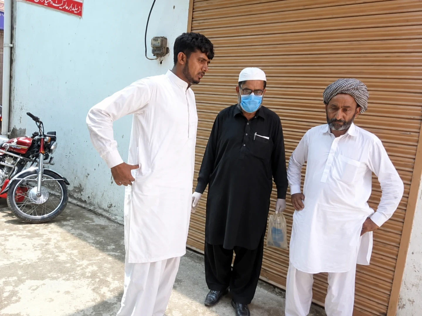 Three men standing outside a shuttered shop; one wears a face mask.