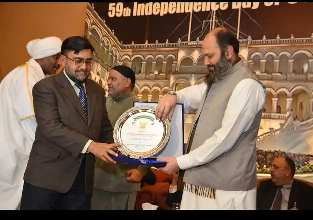 Two men holding a round presentation plaque in front of a formal event backdrop.