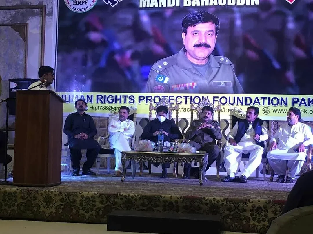 A speaker at a lectern beside seated participants and a large HRPF event screen.
