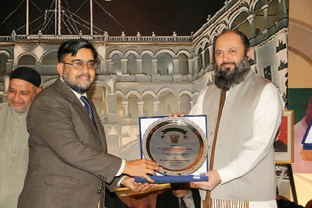 Two men holding a round plaque in front of a printed event backdrop.