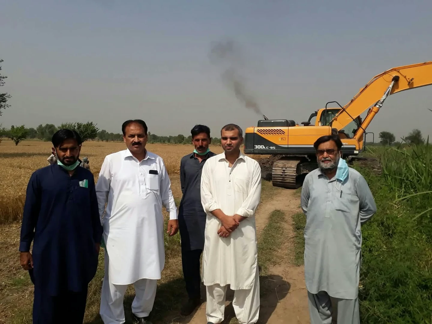 Five men standing in a field with an excavator behind them.