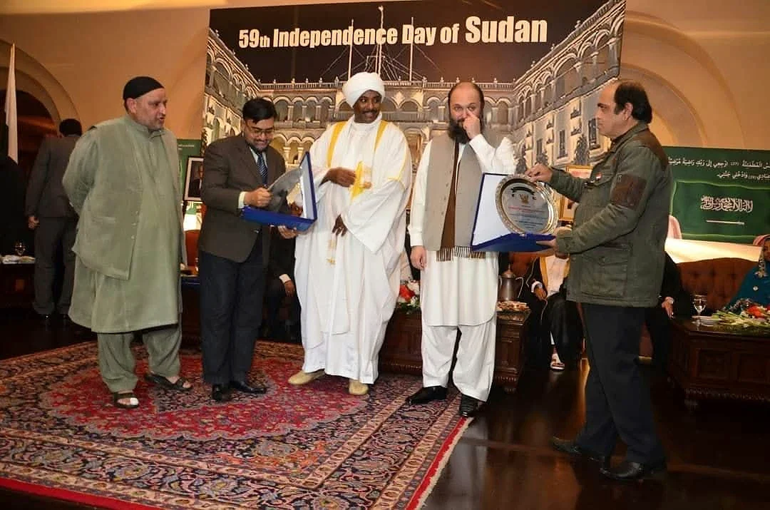 A group posing with presentation plaques in front of a Sudan Independence Day backdrop.