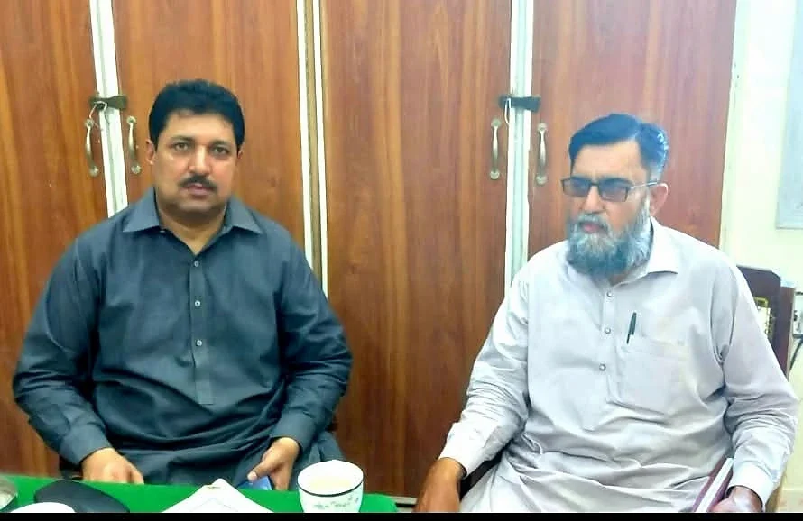 Two men seated at a green meeting table with tea cups in front of them.