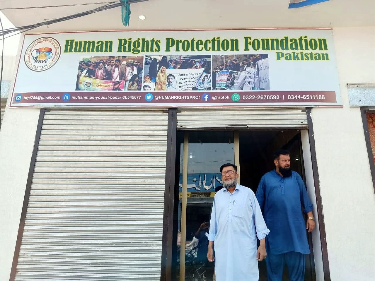 Two men standing beneath a Human Rights Protection Foundation Pakistan sign at an office entrance.