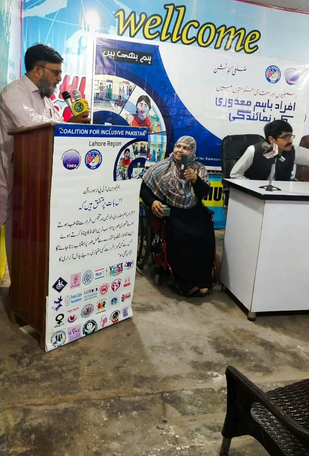 An indoor meeting with a lectern, welcome banner and seated panel.