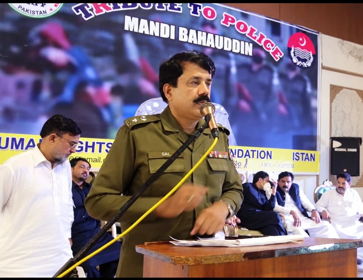 A man in a police uniform speaking into a microphone in front of an HRPF event backdrop.