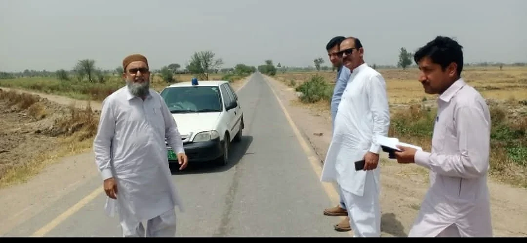 Men standing beside a white car on a rural road bordered by fields.