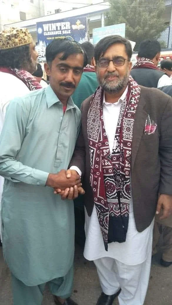 Two men shaking hands at an outdoor gathering; one wears a red patterned shawl.