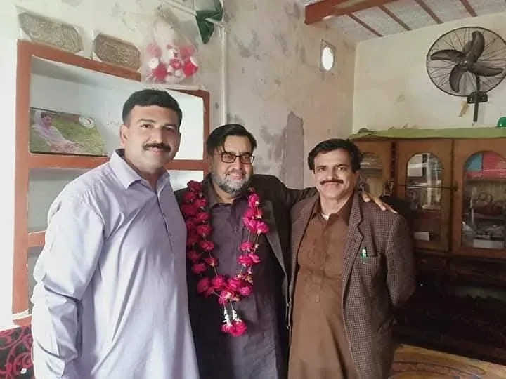 Three men posing indoors, with one wearing flower garlands.