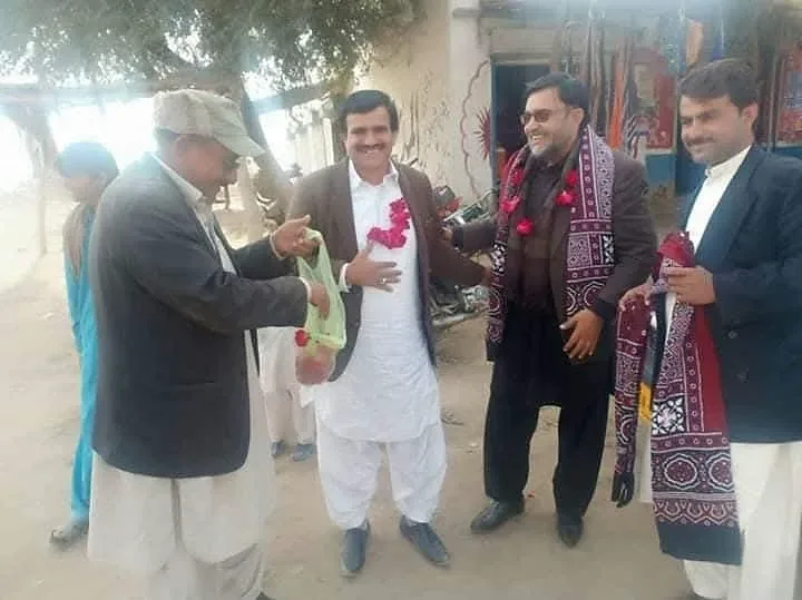 Men exchanging greetings outdoors while wearing flower garlands and patterned shawls.