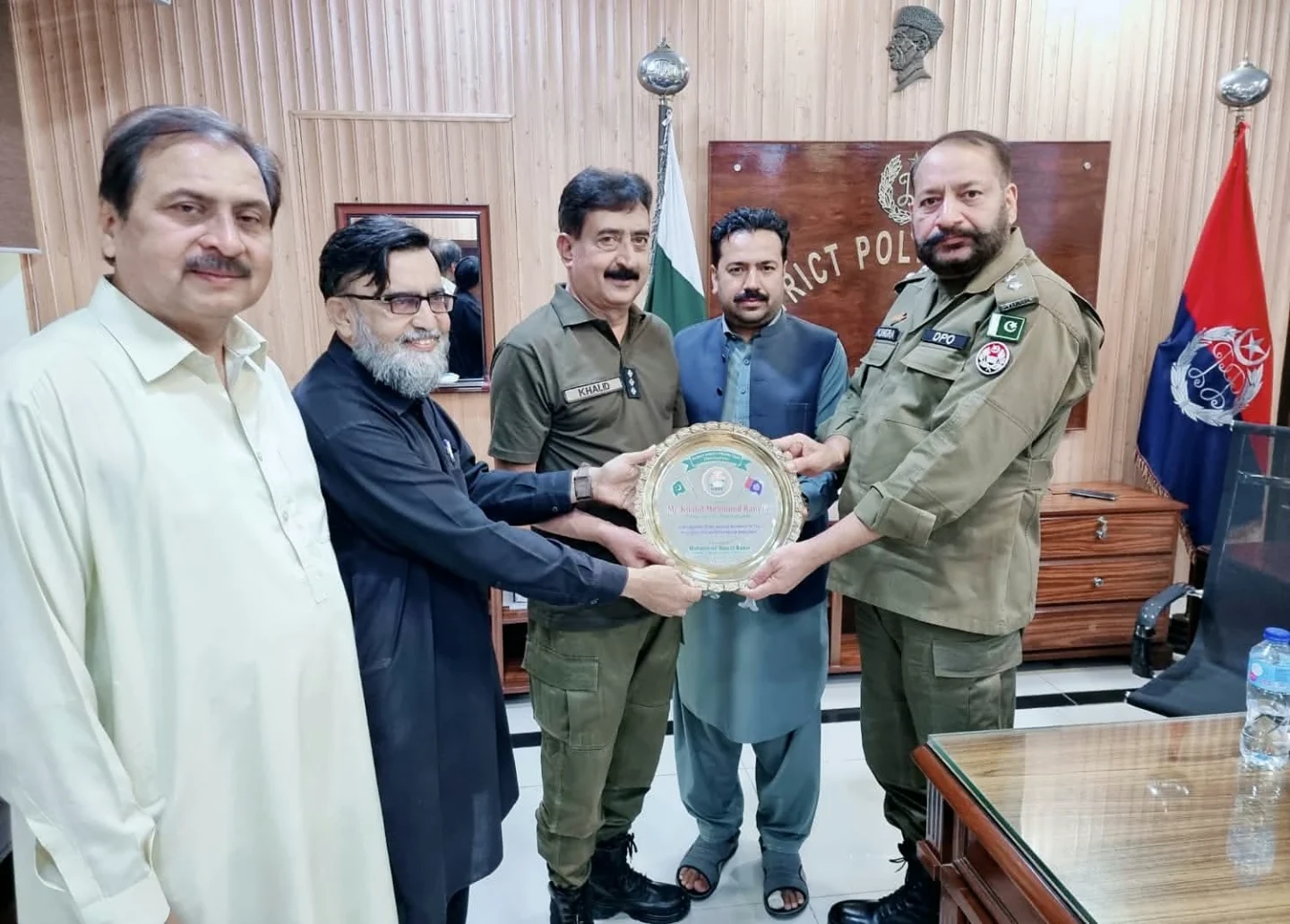 Uniformed personnel and other men holding a round presentation plaque in an office.