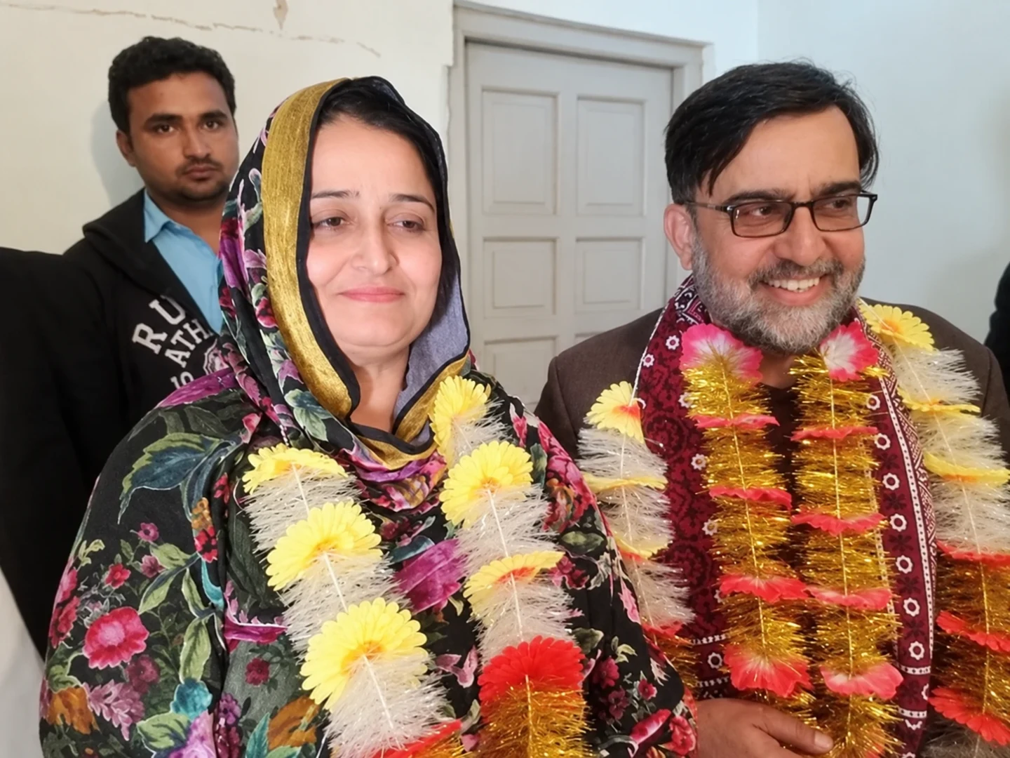 A woman and two men posing indoors, with two wearing bright flower garlands.
