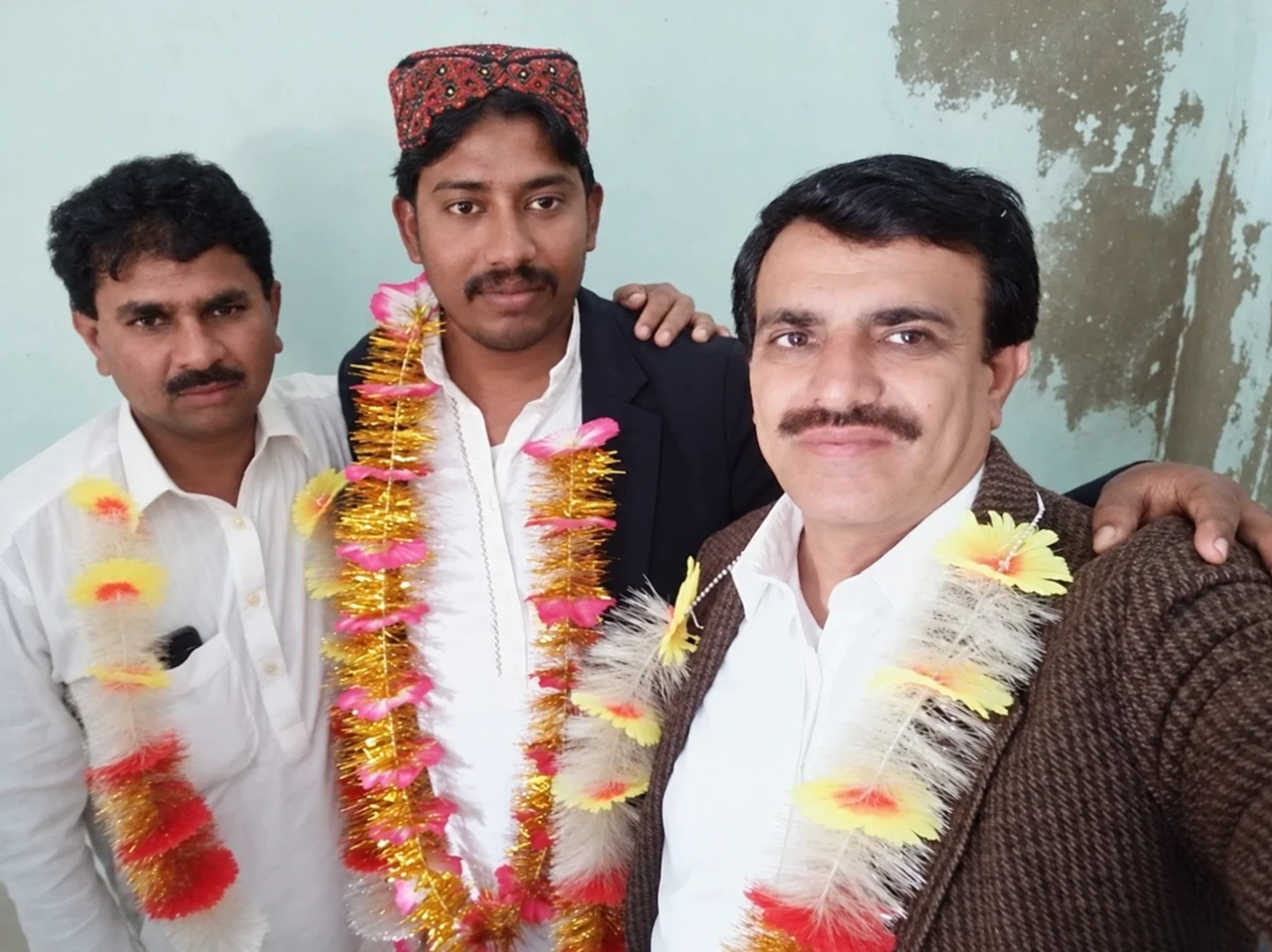 Three men posing together indoors wearing colourful flower garlands.