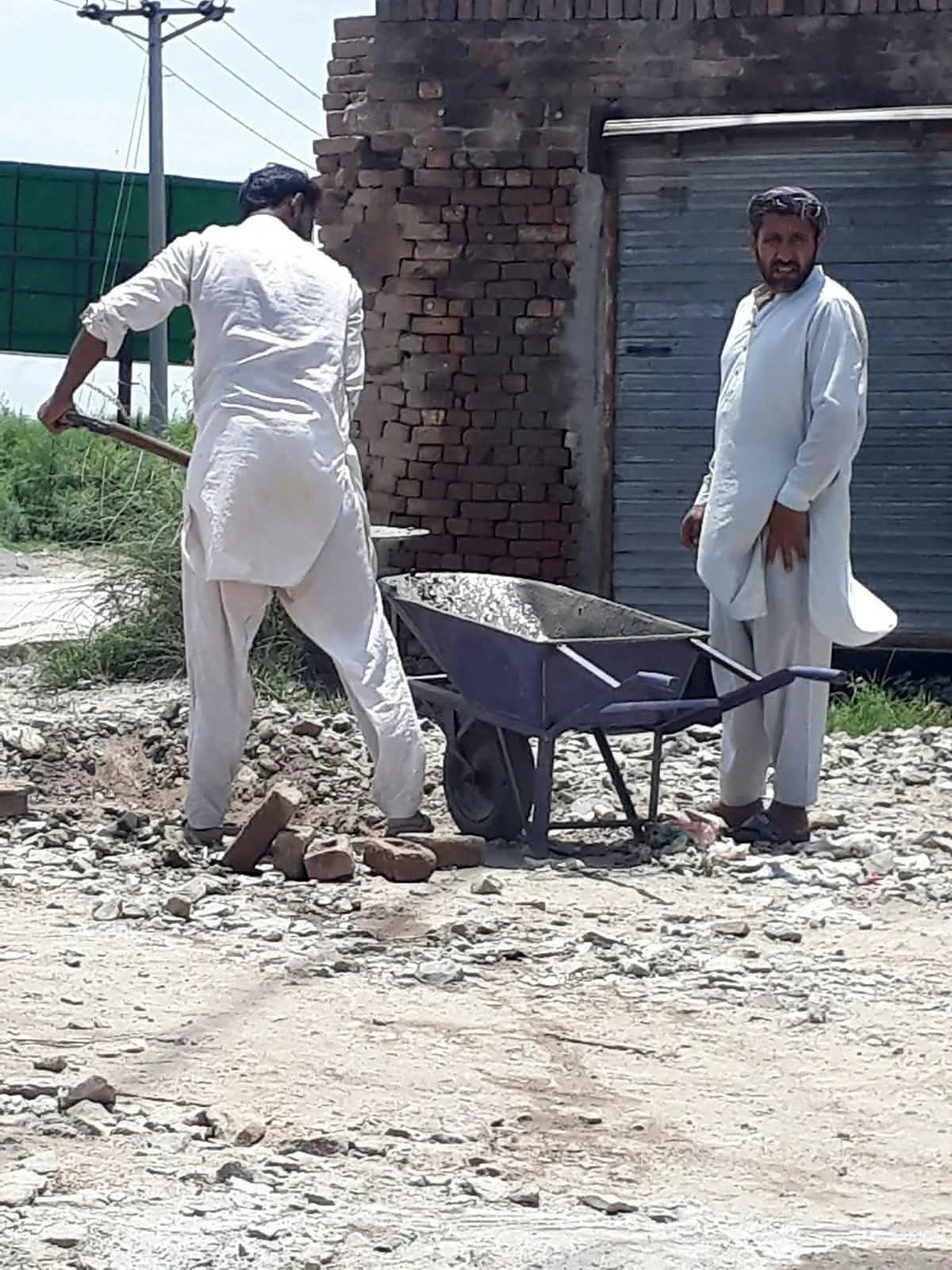 Two men beside a wheelbarrow and a brick building outdoors.