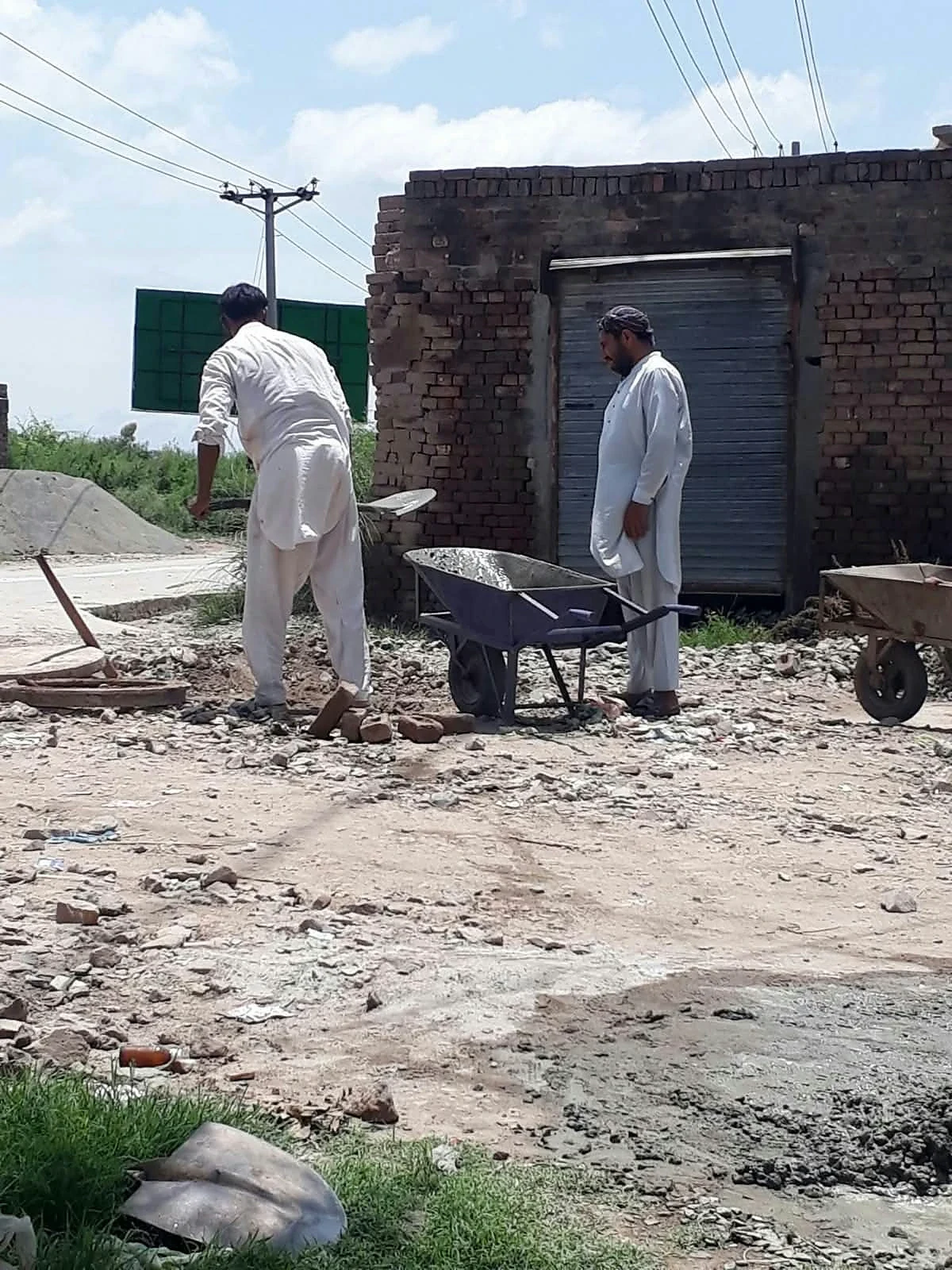 Men standing beside a wheelbarrow on uneven ground outside a brick building.