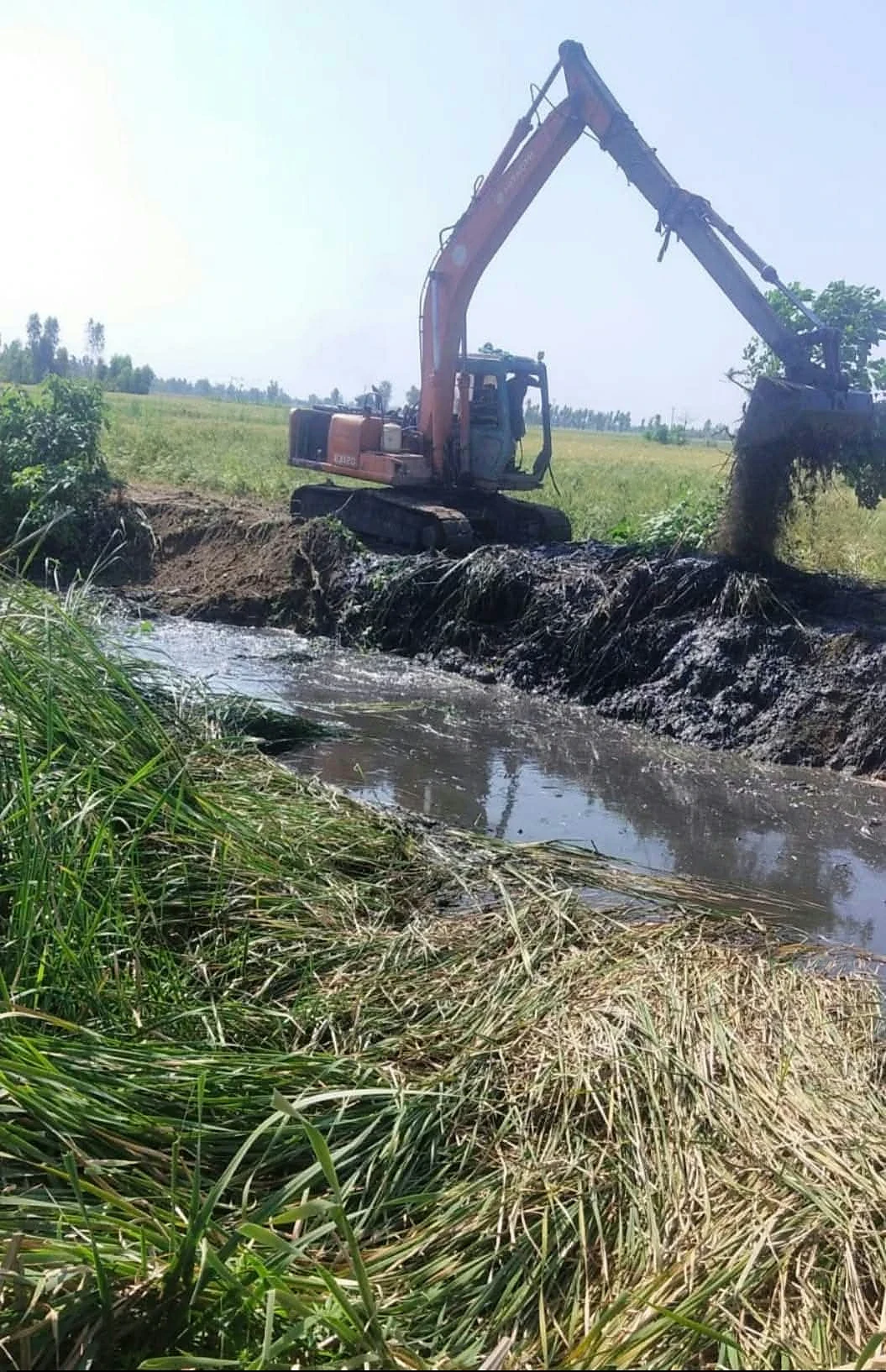 An excavator positioned beside a narrow channel running through fields.