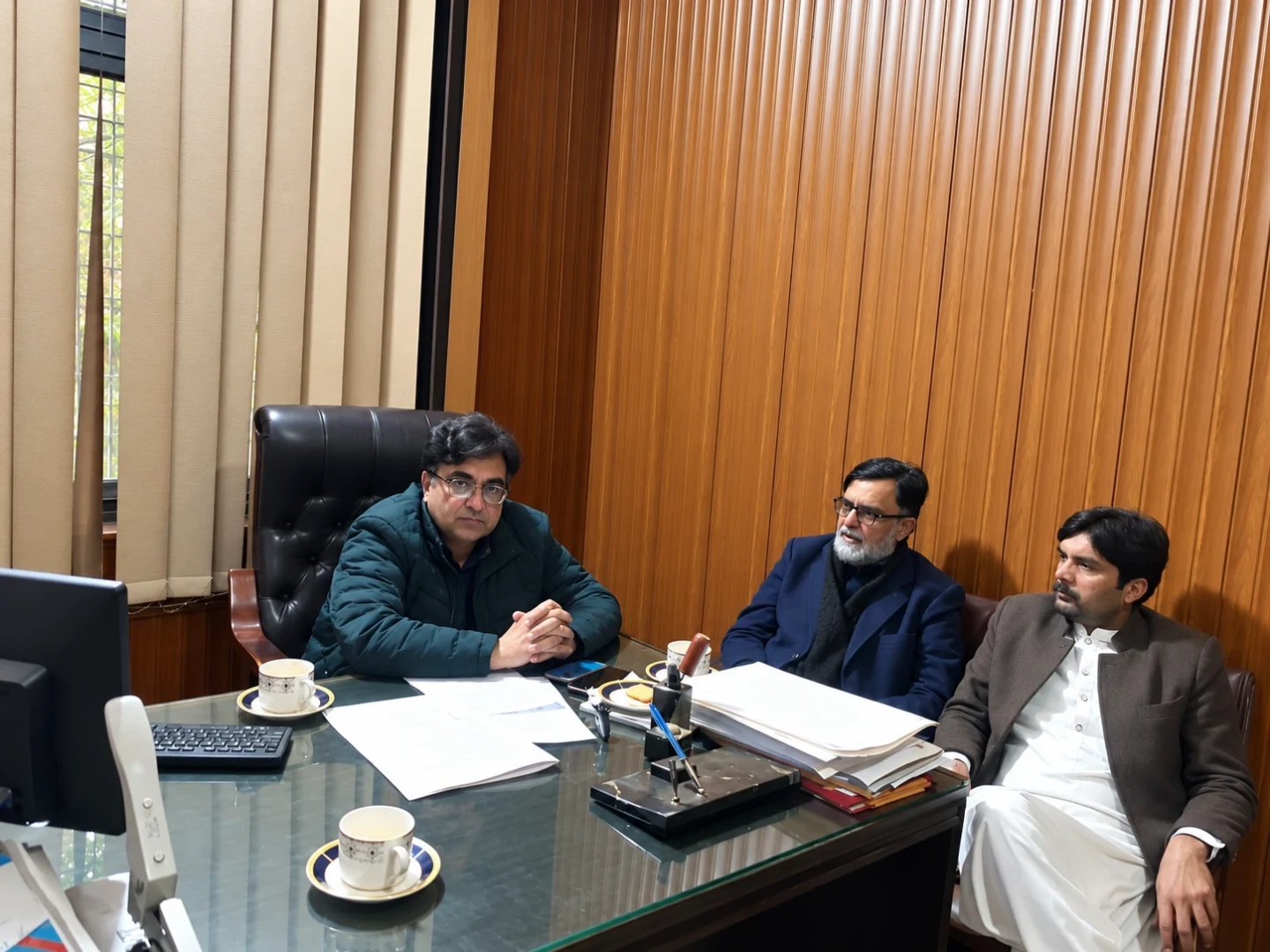 Three men seated around a green office desk with papers and tea cups.