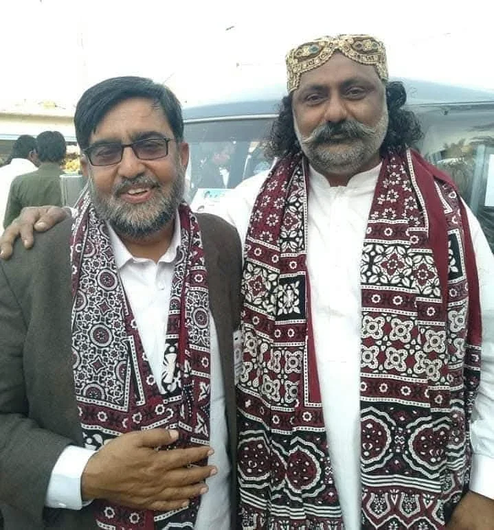 Two men posing outdoors wearing red patterned shawls.