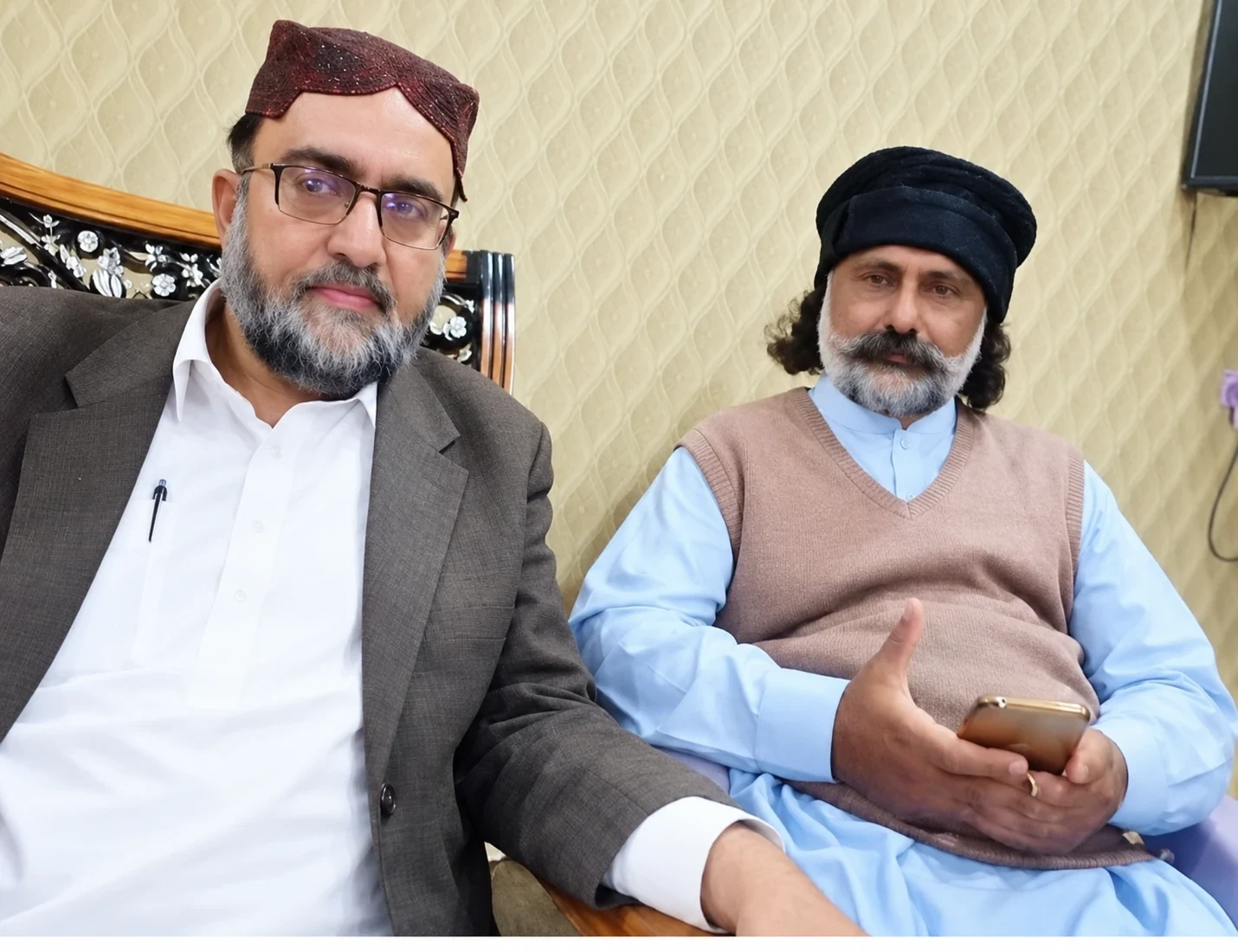 Two men seated together indoors; one wears a black turban.