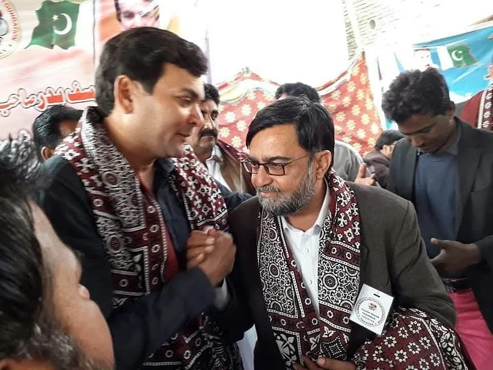 A man wearing garlands and a patterned shawl speaking near microphones in a crowd.