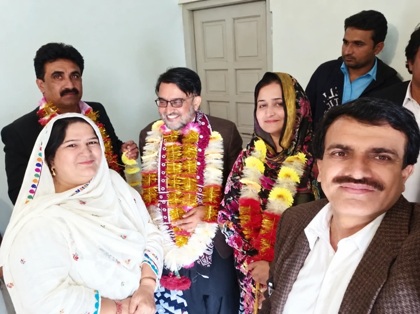 Women and men posing closely together indoors, with flower garlands visible.